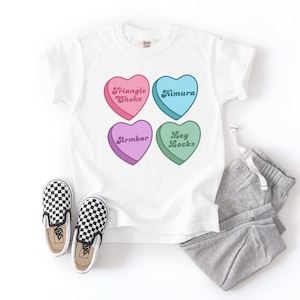 May include: White t-shirt featuring four heart-shaped graphics in pink, blue, purple, and green, each with a different Jiu-Jitsu term. The shirt is paired with grey sweatpants and black and white checkered slip-on shoes.