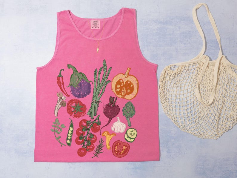 May include: A vibrant pink vest top featuring a vegetable illustration with a red chilli, aubergine, asparagus, and tomatoes. A beige string bag is next to the vest top.
