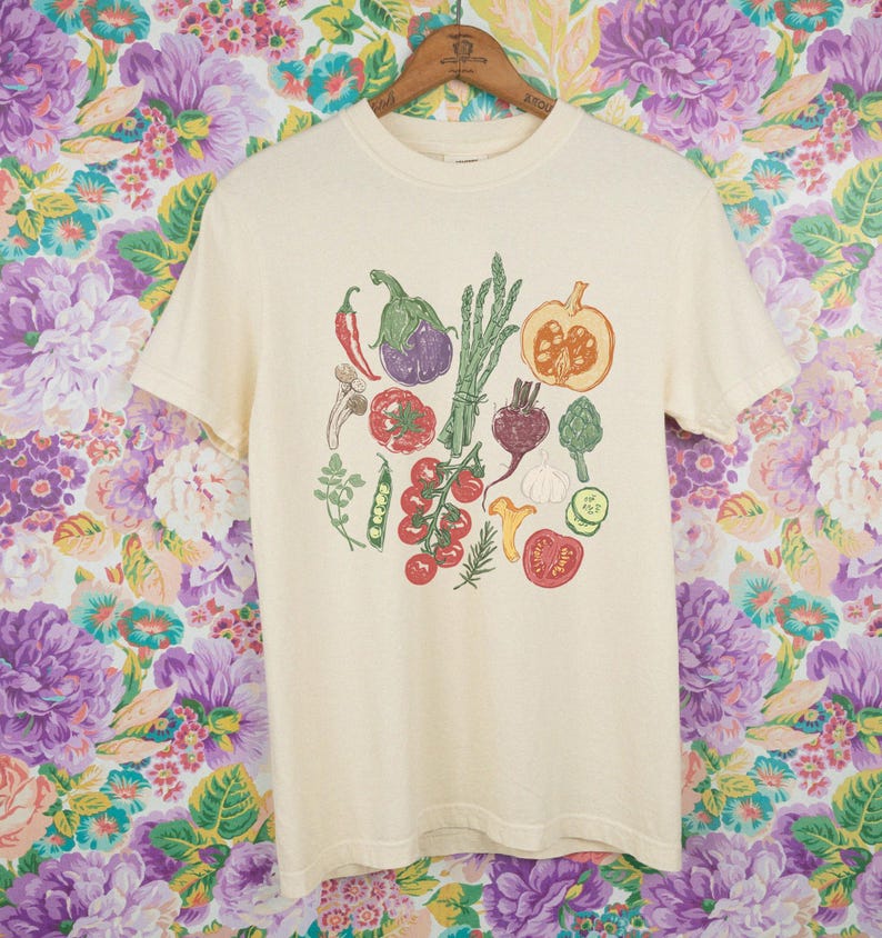 May include: Cream-colored t-shirt featuring a colorful illustration of various vegetables, including tomatoes, eggplant, and asparagus. The design is centered on the front of the shirt. The shirt is hanging on a wooden hanger against a floral background.