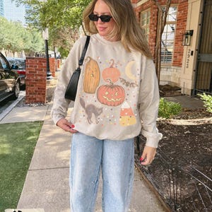 May include: A sand-colored crewneck sweatshirt featuring a graphic print of a tall sad pumpkin, a round jack-o'-lantern, a crescent moon, a flying bat, a black cat, and a piece of candy corn, paired with light blue denim jeans and a black shoulder bag.