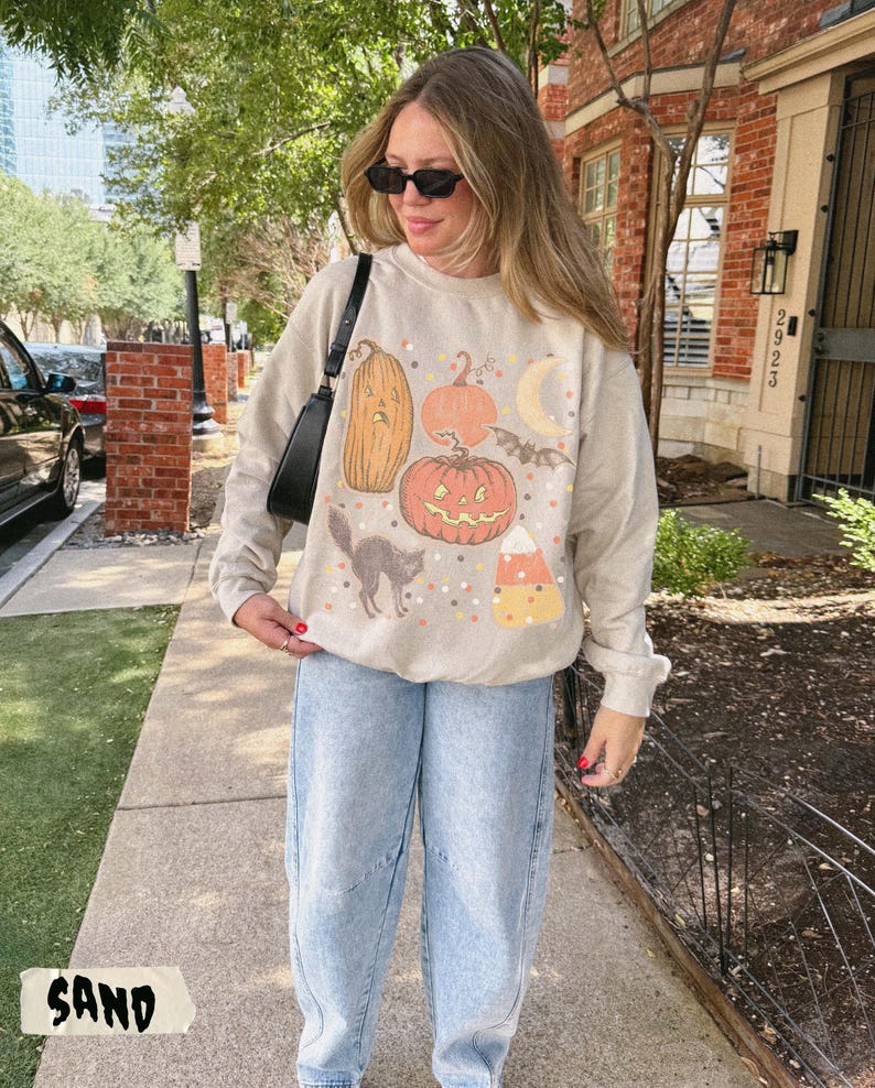 May include: A sand-colored crewneck sweatshirt featuring a graphic print of a tall sad pumpkin, a round jack-o'-lantern, a crescent moon, a flying bat, a black cat, and a piece of candy corn, paired with light blue denim jeans and a black shoulder bag.
