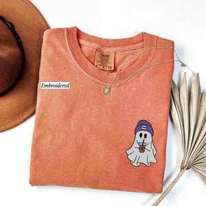May include: Coral-colored t-shirt with a round neck and a small embroidered ghost graphic wearing a purple beanie and holding a coffee cup. A gold necklace and a brown hat are also in the image.