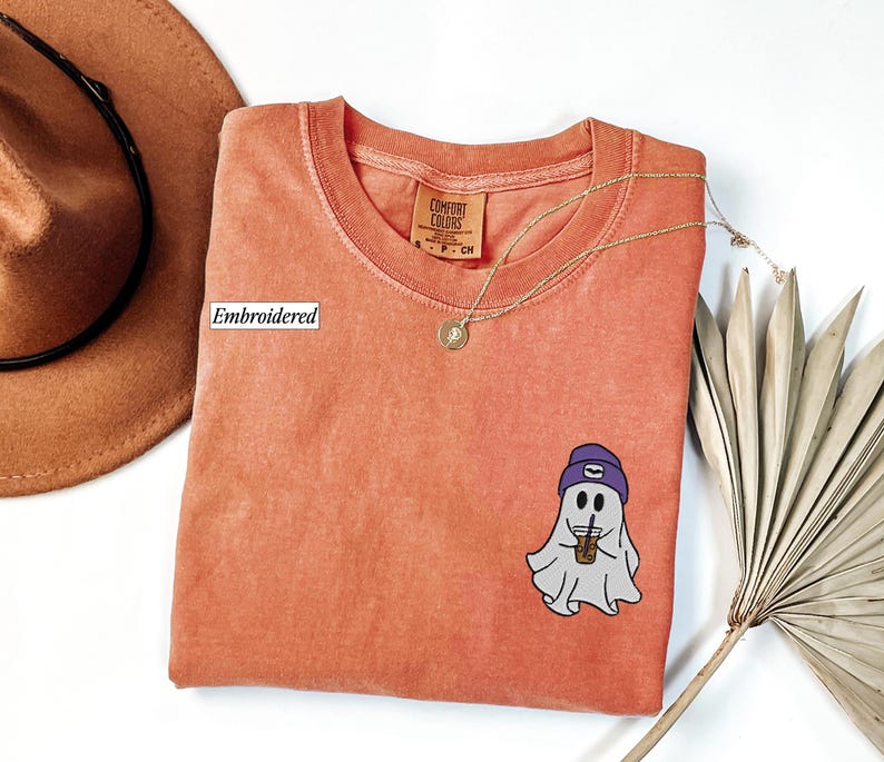 May include: Coral-colored t-shirt with a round neck and a small embroidered ghost graphic wearing a purple beanie and holding a coffee cup. A gold necklace and a brown hat are also in the image.