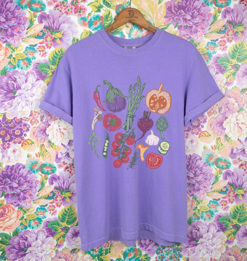 May include: A light purple t-shirt featuring a colorful illustration of various vegetables, including tomatoes, eggplant, and asparagus. The shirt is hanging on a wooden hanger against a floral background.