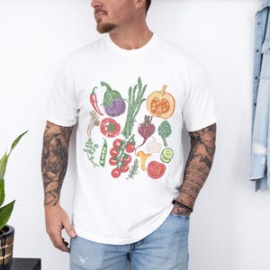 May include: White t-shirt featuring a colorful illustration of various vegetables, including tomatoes, eggplant, asparagus, and a pumpkin. The design is a hand-drawn style with green, red, purple, and orange hues. The shirt is a casual, everyday wear item.