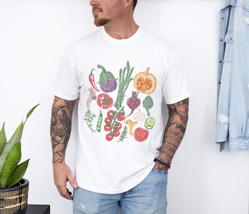 May include: White t-shirt featuring a colorful illustration of various vegetables, including tomatoes, eggplant, asparagus, and a pumpkin. The design is a hand-drawn style with green, red, purple, and orange hues. The shirt is a casual, everyday wear item.