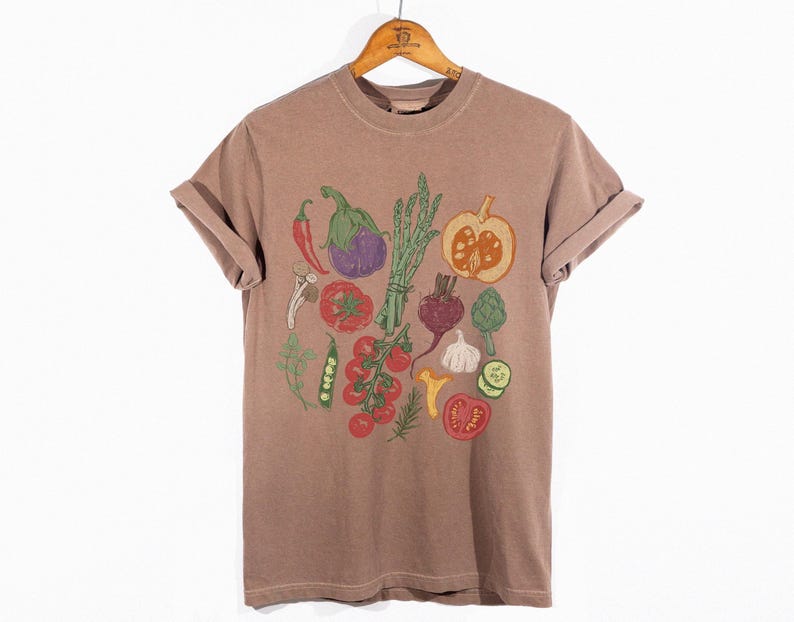 May include: A brown t-shirt with rolled-up sleeves, featuring a colorful illustration of various vegetables. The design includes a red chili pepper, eggplant, asparagus, tomatoes, and a pumpkin, among others. The shirt is hanging on a wooden hanger.