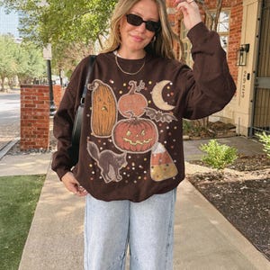 May include: A dark brown long-sleeve crewneck sweatshirt featuring a graphic print of various pumpkins, a black cat, a bat, a crescent moon, and candy corn, accented with scattered multi-colored dots across the front of the garment.