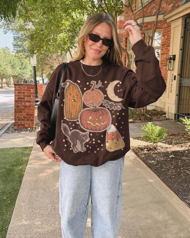 May include: A dark brown long-sleeve crewneck sweatshirt featuring a graphic print of various pumpkins, a black cat, a bat, a crescent moon, and candy corn, accented with scattered multi-colored dots across the front of the garment.