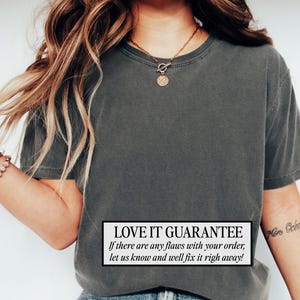 May include: Dark gray t-shirt with a crew neck. The shirt has a slightly faded appearance. A white rectangle at the bottom of the shirt contains the text "LOVE IT GUARANTEE" and additional text. A gold necklace and bracelet are also visible.