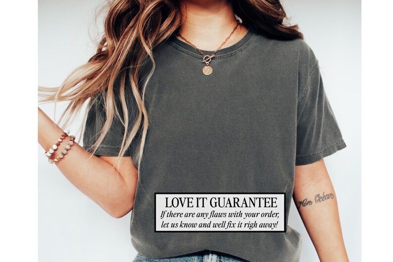 May include: Dark gray t-shirt with a crew neck. The shirt has a slightly faded appearance. A white rectangle at the bottom of the shirt contains the text "LOVE IT GUARANTEE" and additional text. A gold necklace and bracelet are also visible.