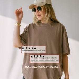 May include: A brown t-shirt with the text "ORIGINAL DESIGNS BY SELLER" and customer reviews. The shirt is worn by a person wearing sunglasses and a cap. The reviews say "Wonderful embroidery. Thank You!" and "This shirt is amazing, and even better it's printed on a Comfort Colors t-shirt (my fav) <3".