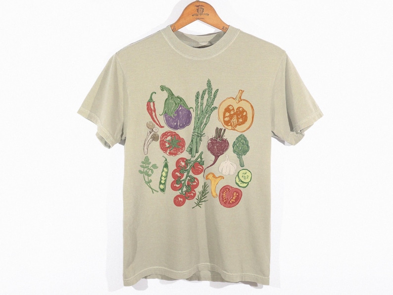 May include: A light tan t-shirt featuring a colorful illustration of various vegetables. The design includes a red chili pepper, eggplant, asparagus, pumpkin, tomatoes, and more. The shirt is hanging on a wooden hanger.