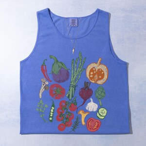 May include: A blue vest top with a vibrant illustration of vegetables. The design includes a red chilli, aubergine, asparagus, tomatoes, and a pumpkin. The top has a scoop neck and a tag at the top.