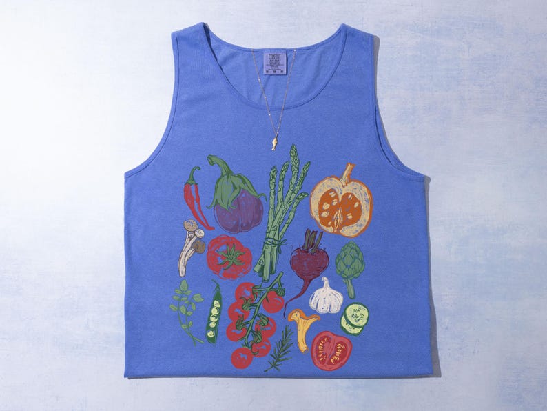 May include: A blue vest top with a vibrant illustration of vegetables. The design includes a red chilli, aubergine, asparagus, tomatoes, and a pumpkin. The top has a scoop neck and a tag at the top.