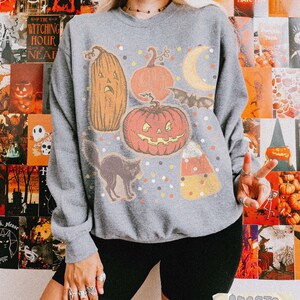 May include: A heather grey crewneck sweatshirt featuring a graphic print of a tall jack-o'-lantern, a round jack-o'-lantern, a crescent moon, a bat, a black cat, and a piece of candy corn, all surrounded by small colorful confetti-like dots.