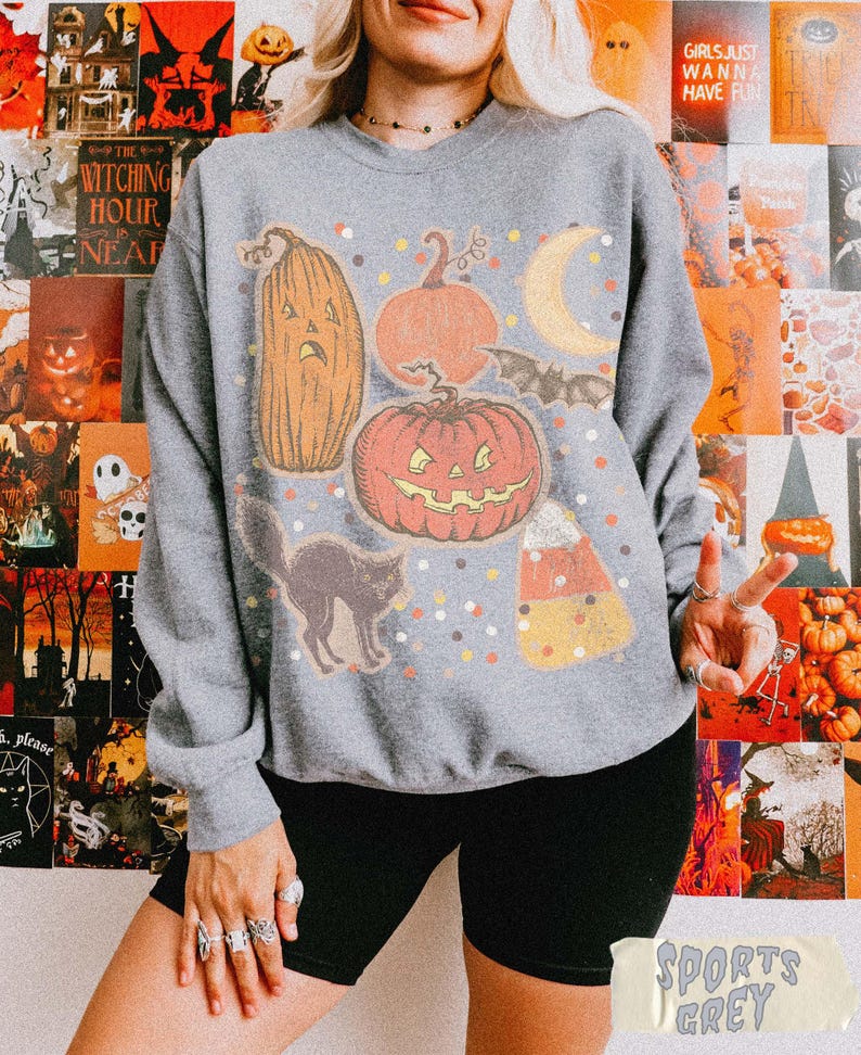 May include: A heather grey crewneck sweatshirt featuring a graphic print of a tall jack-o'-lantern, a round jack-o'-lantern, a crescent moon, a bat, a black cat, and a piece of candy corn, all surrounded by small colorful confetti-like dots.