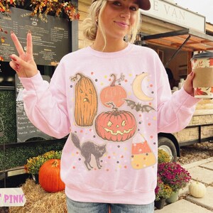 May include: A light pink crewneck sweatshirt featuring a graphic print of various Halloween-themed illustrations, including a tall pumpkin, a round jack-o'-lantern, a crescent moon, a flying bat, a black cat, and a piece of candy corn.