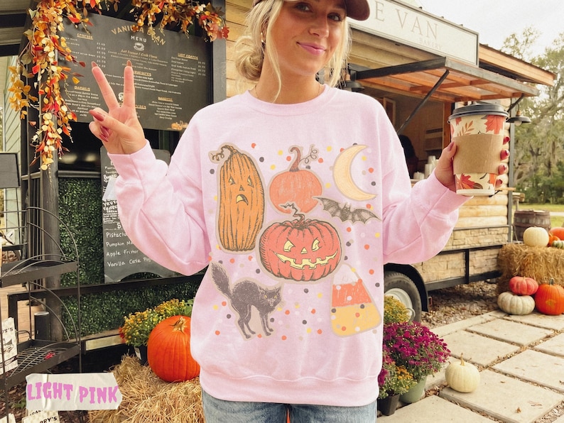 May include: A light pink crewneck sweatshirt featuring a graphic print of various Halloween-themed illustrations, including a tall pumpkin, a round jack-o'-lantern, a crescent moon, a flying bat, a black cat, and a piece of candy corn.
