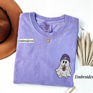 May include: A lavender Comfort Colors t-shirt with an embroidered ghost wearing a purple beanie and holding an iced coffee. The shirt has a crew neck and a small tag. A gold necklace and a brown hat are also in the image.