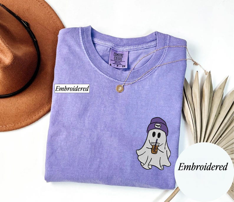 May include: A lavender Comfort Colors t-shirt with an embroidered ghost wearing a purple beanie and holding an iced coffee. The shirt has a crew neck and a small tag. A gold necklace and a brown hat are also in the image.
