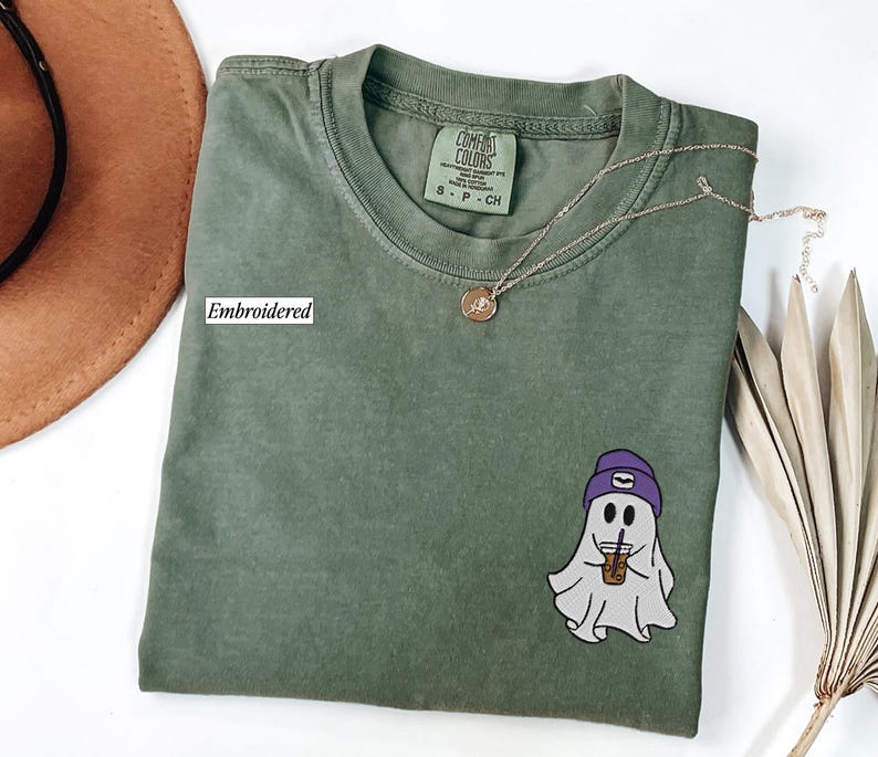 May include: A green Comfort Colors t-shirt with an embroidered ghost graphic. The ghost wears a purple beanie and holds a coffee cup. The shirt has a Comfort Colors tag and a gold necklace. The word 'Embroidered' is printed on the shirt.