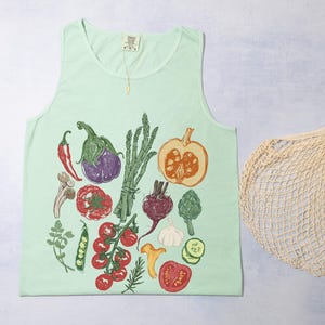 May include: A pale green vest top with a vibrant illustration of vegetables such as aubergine, tomatoes, and a pumpkin. A beige mesh shopping bag is on the right side of the vest top.