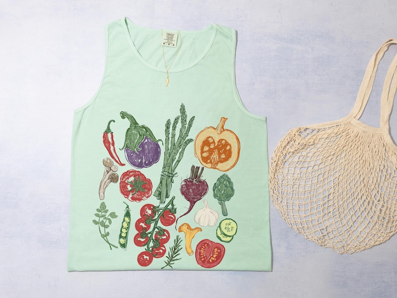 May include: A pale green vest top with a vibrant illustration of vegetables such as aubergine, tomatoes, and a pumpkin. A beige mesh shopping bag is on the right side of the vest top.