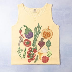 May include: A pale yellow vest top with a colourful print of vegetables such as aubergine, tomatoes, and asparagus. The design has a hand-drawn style, suitable for a farmer's market or anyone who enjoys gardening. The print includes a pumpkin.