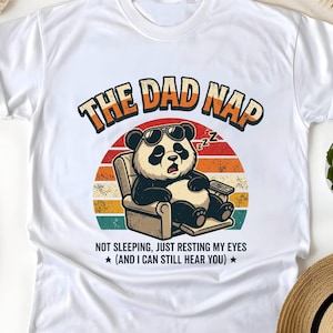 Może przedstawiać: Biała koszulka z pandą w okularach przeciwsłonecznych, relaksującą się na krześle z pilotem. Koszulka ma napis "THE DAD NAP" nad pandą i "NOT SLEEPING, JUST RESTING MY EYES (AND I CAN STILL HEAR YOU)" poniżej.