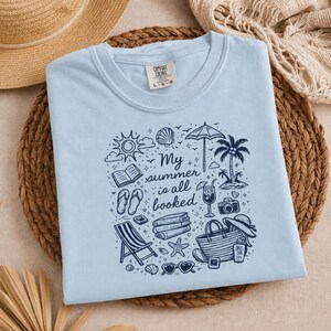 May include: A light blue t-shirt featuring a dark blue graphic print with the text "My summer is all booked" surrounded by illustrations of a sun, books, flip-flops, a beach chair, a palm tree, a cocktail, a camera, a tote bag, and sunglasses.