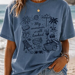 May include: A blue short-sleeved t-shirt featuring a dark blue graphic print of summer holiday items including a beach umbrella, palm tree, books, flip-flops, a tote bag, and sunglasses, with the text My summer is all booked in the centre.