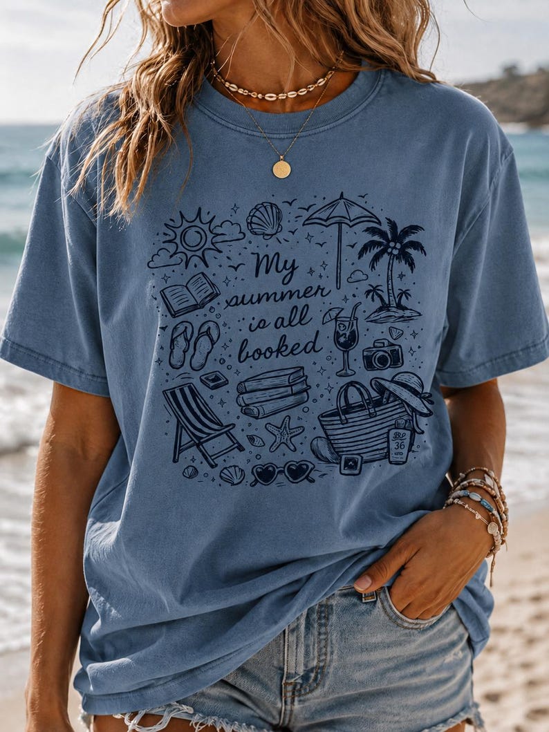 May include: A blue short-sleeved t-shirt featuring a dark blue graphic print of summer holiday items including a beach umbrella, palm tree, books, flip-flops, a tote bag, and sunglasses, with the text My summer is all booked in the centre.