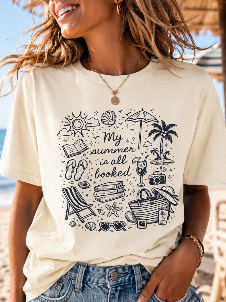 May include: A cream-coloured t-shirt featuring a black graphic print of summer-themed icons including a sun, books, flip-flops, a beach chair, a palm tree, a cocktail, a camera, a tote bag, and sunglasses with the text My summer is all booked.