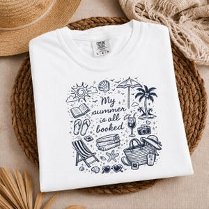May include: A white t-shirt featuring a black ink graphic design with the text My summer is all booked. The illustration includes a sun, seashell, umbrella, palm tree, open book, flip-flops, stack of books, cocktail, camera, beach bag, and sunglasses.