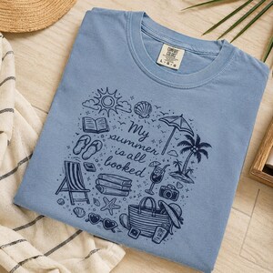 May include: A dusty blue t-shirt featuring a dark blue graphic print with the text "My summer is all booked" surrounded by illustrations of a sun, a beach umbrella, a palm tree, books, a beach chair, sandals, sunglasses, and a tote bag.
