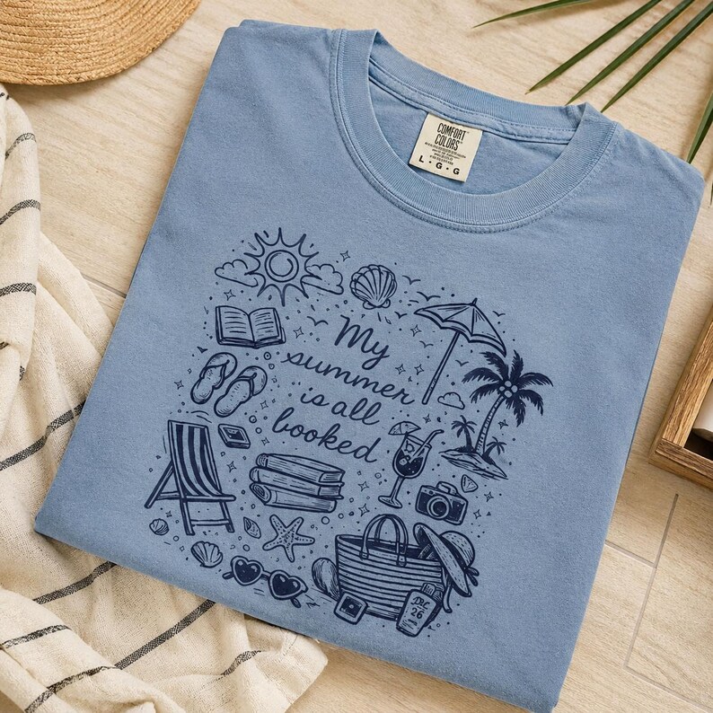 May include: A dusty blue t-shirt featuring a dark blue graphic print with the text "My summer is all booked" surrounded by illustrations of a sun, a beach umbrella, a palm tree, books, a beach chair, sandals, sunglasses, and a tote bag.