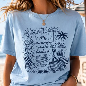 May include: A light blue t-shirt featuring a dark blue graphic print of summer holiday items including a beach umbrella, palm tree, books, flip-flops, a tote bag, and sunglasses, with the text "My summer is all booked" in the centre.