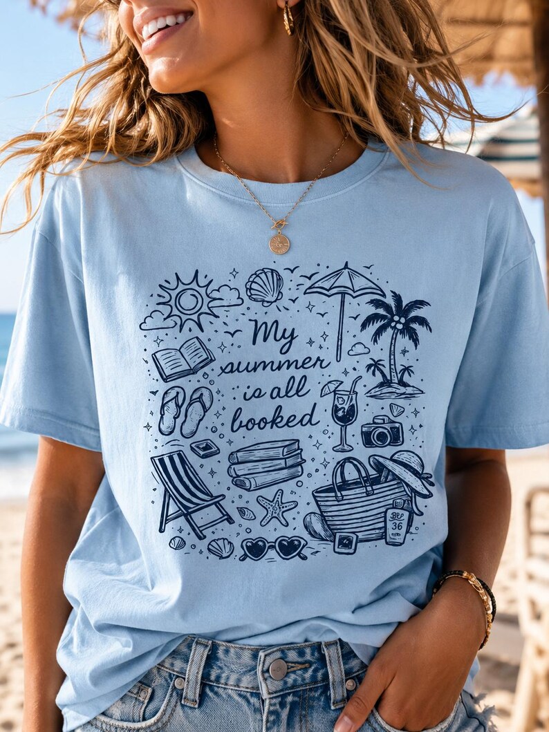 May include: A light blue t-shirt featuring a dark blue graphic print of summer holiday items including a beach umbrella, palm tree, books, flip-flops, a tote bag, and sunglasses, with the text "My summer is all booked" in the centre.