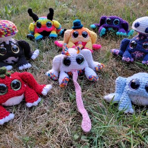 May include: A collection of eight crocheted stuffed toys, including a mushroom, a strawberry, a frog, a spider, a unicorn, and a monster. The toys are all brightly colored and have large, expressive eyes.