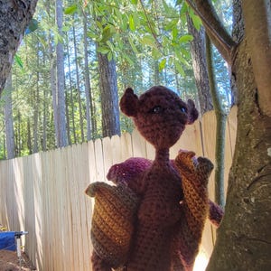 Puede incluir: Un juguete de peluche tejido a crochet con un cuerpo marrón y orejas grandes. El juguete está de pie frente a una valla de madera y un árbol.