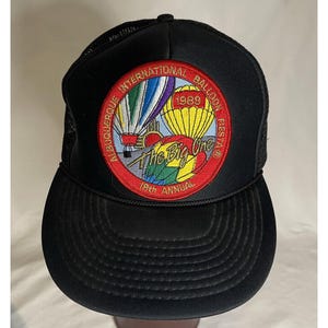 May include: Black mesh trucker hat with a red, white and blue embroidered patch that says "Albuquerque International Balloon Fiesta 1989 The Big One 18th Annual".