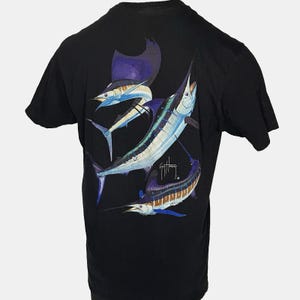 May include: Black t-shirt with a vibrant illustration of three sailfish in shades of blue, white, and brown. The artist's signature is visible. The t-shirt has a crew neck design, a classic style for casual wear.