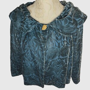 May include: A teal blue velvet jacket with a large, wide collar. The jacket has a floral pattern in a darker shade of blue. A gold leaf-shaped button is visible on the front of the jacket.