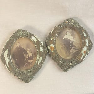 Könnte beinhalten: Zwei ovale antike Bilderrahmen mit Sepia-Porträts. Die verzierten Rahmen haben ein abgenutztes, gealtertes Aussehen mit abblätternder weißer Farbe auf einer metallischen Basis. Einer zeigt ein Paar, der andere ein Kind.