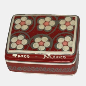 May include: A red ceramic trinket box with a floral design. The box has a white border and a gray floral pattern. The words "Taxco - Mexico" are written on the front of the box.