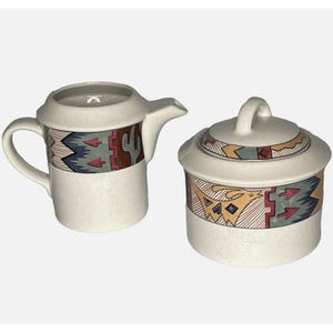May include: A cream-colored ceramic creamer and sugar set with a southwestern design. The creamer has a handle and a spout. The sugar bowl has a lid. Both pieces feature a geometric pattern in shades of blue, green, red, and brown.