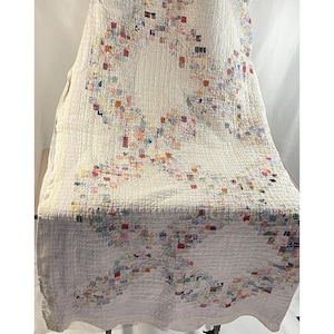 May include: A vintage white quilt with a repeating pattern of colorful, small square patches. The quilt has a quilted texture and a large, open heart-shaped design in the center. The patches are in various colors, including red, blue, yellow, and green.