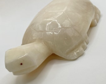 May include: A carved, off-white turtle figurine. The turtle has a detailed shell pattern and a small red dot for an eye. The figurine is made of a smooth, possibly stone-like material.