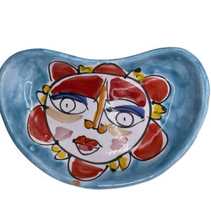 May include: A hand-painted ceramic serving dish with a whimsical face design. The dish has a light blue background and a face with red, yellow, and white accents. The face has blue eyes and red lips. The dish is oval-shaped.